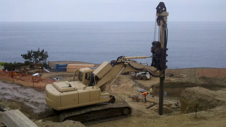 A large tracked drilling machine operates on a sandy construction site near the ocean, with building materials and a worker visible in the background.