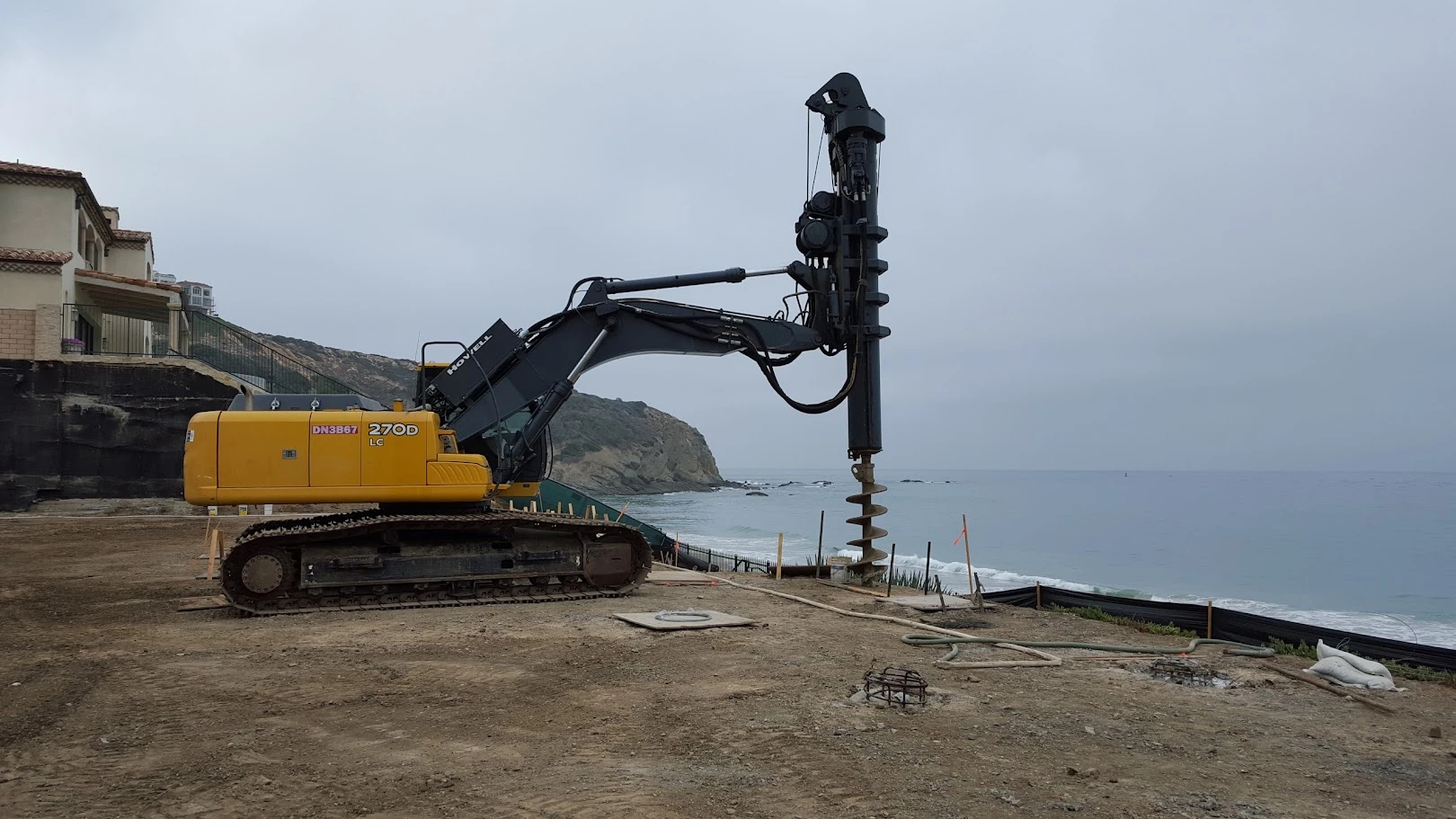 A large yellow drilling machine with an auger attachment is stationed on bare ground near a coastline, with the ocean and cloudy sky in the background.