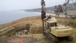 A large drilling machine operates on a coastal construction site overlooking the ocean, with fencing and excavated earth visible in the background.