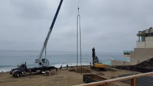 A construction crane and excavator operate on a sandy beach near the ocean, with building materials and equipment scattered around the site.