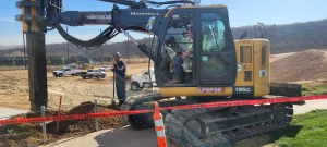 A construction worker stands next to a large drilling excavator behind a "danger" tape at a construction site with vehicles and dirt piles in the background.
