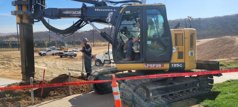 A construction worker stands next to a large drilling excavator behind a "danger" tape at a construction site with vehicles and dirt piles in the background.