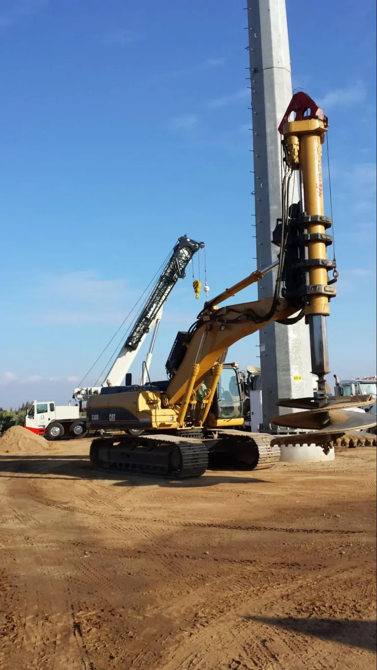 A large yellow drilling machine is installing a concrete pile into the ground at a construction site, with a crane and other equipment in the background.
