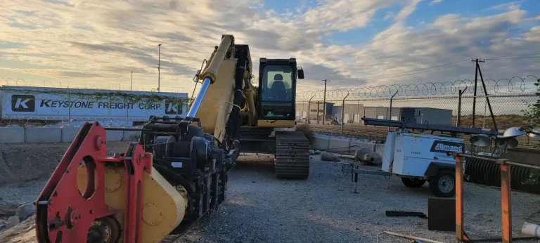 A yellow excavator is parked at a gravel construction site near a trailer and equipment, with a "Keystone Freight Corp" banner on the fence in the background.