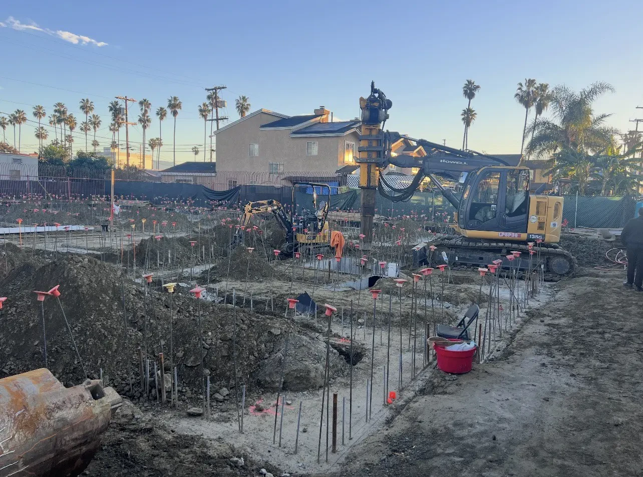 Construction site with exposed rebar, excavators, and workers preparing foundations; houses and palm trees are visible in the background under a clear sky.