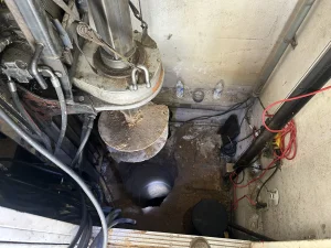 A close-up view of a drilling rig positioned over a deep, cylindrical hole in the ground, surrounded by cables, pipes, and a few water bottles against a wall.