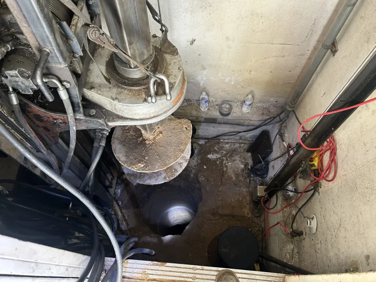 A close-up view of a drilling rig positioned over a deep, cylindrical hole in the ground, surrounded by cables, pipes, and a few water bottles against a wall.