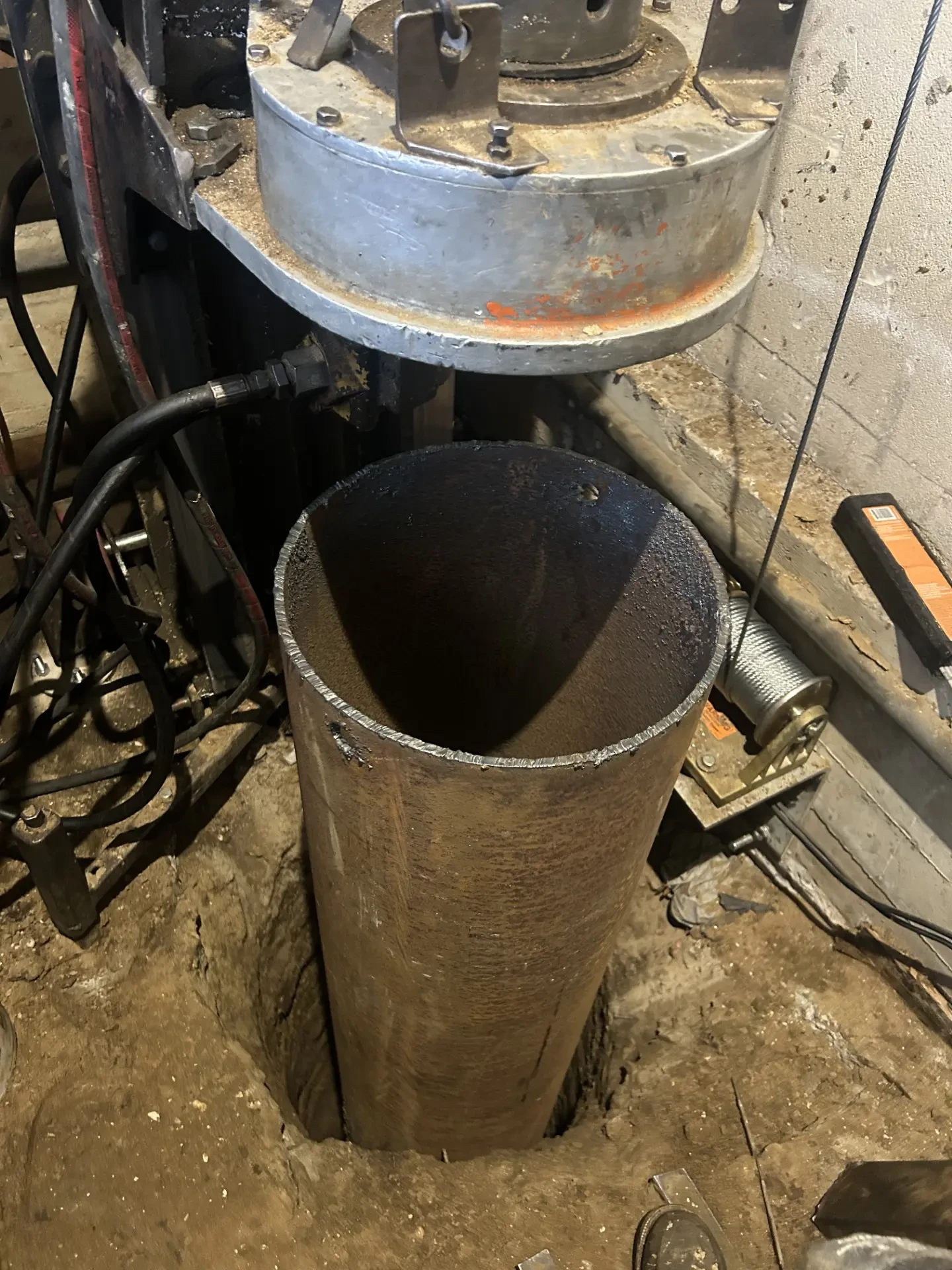 Large metal pipe vertically inserted into a hole in the ground, surrounded by machinery, cables, and a concrete floor in an industrial setting.